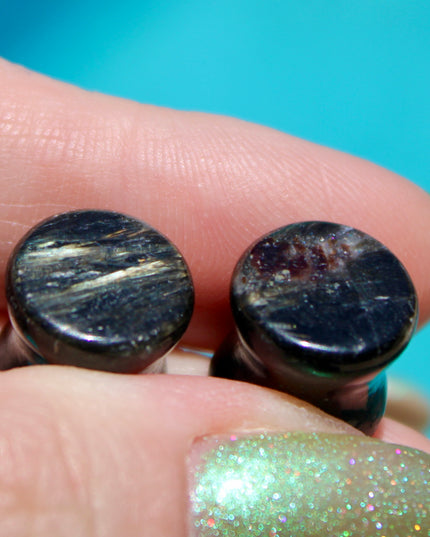 A stunning pair of Astrophyllite FLASH Double Flare Plugs rests against a dark, textured backdrop, their surfaces gleaming with bursts of golden-bronze metallic flash. Each plug is crafted from genuine astrophyllite, a rare and complex stone known for its radiant, starburst-like inclusions that shimmer brilliantly under light. The deep black and charcoal base is layered with reflective veins and flashes of copper, gold, and occasionally blue—giving these plugs a cosmic, otherworldly appearance.