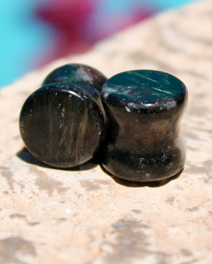 A stunning pair of Astrophyllite FLASH Double Flare Plugs rests against a dark, textured backdrop, their surfaces gleaming with bursts of golden-bronze metallic flash. Each plug is crafted from genuine astrophyllite, a rare and complex stone known for its radiant, starburst-like inclusions that shimmer brilliantly under light. The deep black and charcoal base is layered with reflective veins and flashes of copper, gold, and occasionally blue—giving these plugs a cosmic, otherworldly appearance.