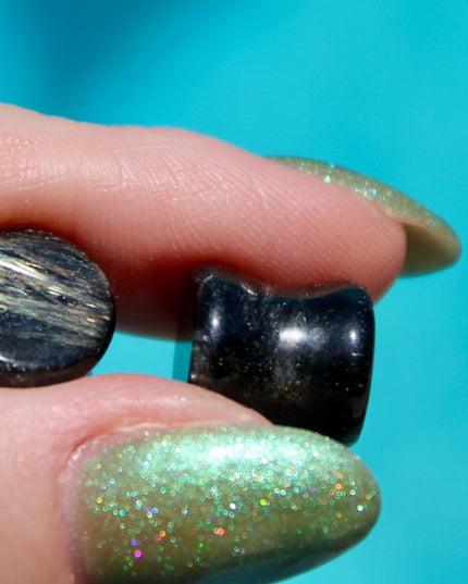 A stunning pair of Astrophyllite FLASH Double Flare Plugs rests against a dark, textured backdrop, their surfaces gleaming with bursts of golden-bronze metallic flash. Each plug is crafted from genuine astrophyllite, a rare and complex stone known for its radiant, starburst-like inclusions that shimmer brilliantly under light. The deep black and charcoal base is layered with reflective veins and flashes of copper, gold, and occasionally blue—giving these plugs a cosmic, otherworldly appearance.