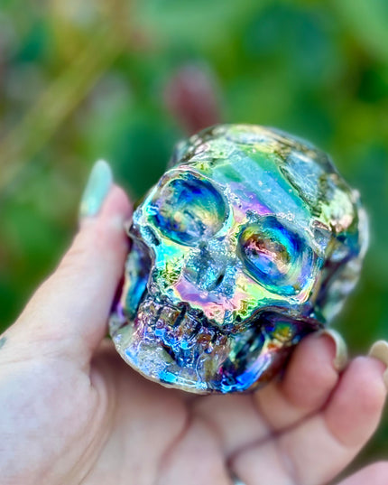 A mesmerizing Genuine Quartz Skull Carving with Rainbow Aura Treatment glows with an ethereal spectrum of colors, resting against a dark, reflective surface. The quartz base is expertly carved into a detailed skull, complete with defined features and a polished finish. The surface shimmers with iridescent hues—vivid blues, purples, pinks, and golds—created through the rainbow aura treatment, a process that bonds precious metals to the quartz for a permanent, luminous effect.