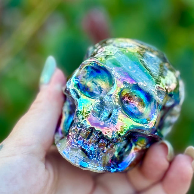 A mesmerizing Genuine Quartz Skull Carving with Rainbow Aura Treatment glows with an ethereal spectrum of colors, resting against a dark, reflective surface. The quartz base is expertly carved into a detailed skull, complete with defined features and a polished finish. The surface shimmers with iridescent hues—vivid blues, purples, pinks, and golds—created through the rainbow aura treatment, a process that bonds precious metals to the quartz for a permanent, luminous effect.