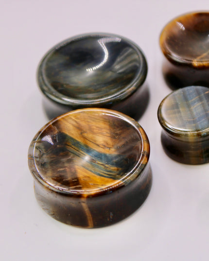 A sleek and polished pair of Blue Tiger Eye plugs, also known as Falcon’s Eye, radiating deep shades of midnight blue, slate, and hints of black. Each plug features the signature chatoyancy of the stone—a silky, light-catching shimmer that moves across the surface as it shifts in the light. The smooth, double-flared design offers both comfort and style, with each plug showcasing natural banding and a unique pattern.