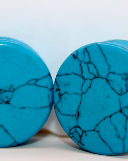A vibrant pair of Blue Howlite Stone Plugs is shown resting on a soft beige surface. The plugs are round, polished to a high shine, and display a brilliant turquoise-blue color. Each plug is marked with distinctive dark gray to black veining in a web-like pattern—natural to howlite that has been dyed blue to resemble turquoise. The smooth, glossy finish enhances the contrast between the bright blue background and the delicate marbling. The lighting gently reflects off the curved surfaces, emphasizing their