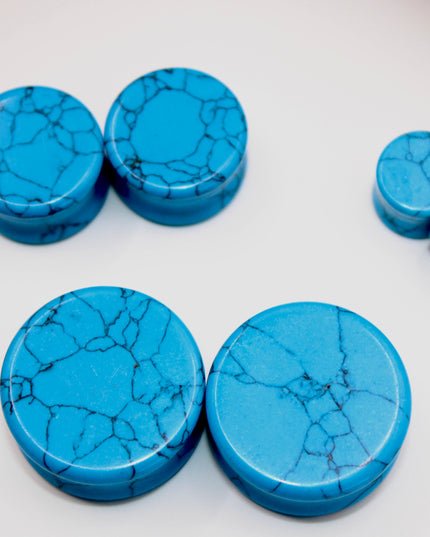 A vibrant pair of Blue Howlite Stone Plugs is shown resting on a soft beige surface. The plugs are round, polished to a high shine, and display a brilliant turquoise-blue color. Each plug is marked with distinctive dark gray to black veining in a web-like pattern—natural to howlite that has been dyed blue to resemble turquoise. The smooth, glossy finish enhances the contrast between the bright blue background and the delicate marbling. The lighting gently reflects off the curved surfaces, emphasizing their
