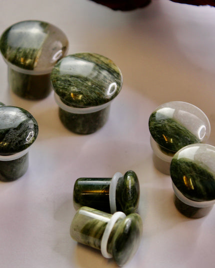 A striking pair of Green Jasper Single Flare Plugs is displayed against an earthy-toned backdrop. Each plug features deep forest green hues with natural patterns of dark veining and subtle swirls, unique to every piece. The surface is smoothly polished, bringing out the rich, organic tones of the stone. Designed with a single flare and a comfortable wearable area, they include O-rings for a secure fit.