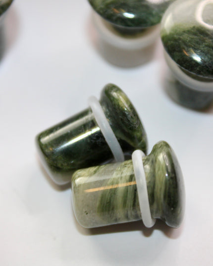 A striking pair of Green Jasper Single Flare Plugs is displayed against an earthy-toned backdrop. Each plug features deep forest green hues with natural patterns of dark veining and subtle swirls, unique to every piece. The surface is smoothly polished, bringing out the rich, organic tones of the stone. Designed with a single flare and a comfortable wearable area, they include O-rings for a secure fit.