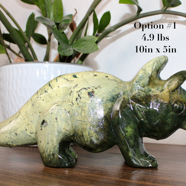 Green stone triceratops figurine on a wooden surface with plants in the background