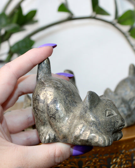 Hand holding a small stone Pyrite kitty cat carving another similar figurine in the background.