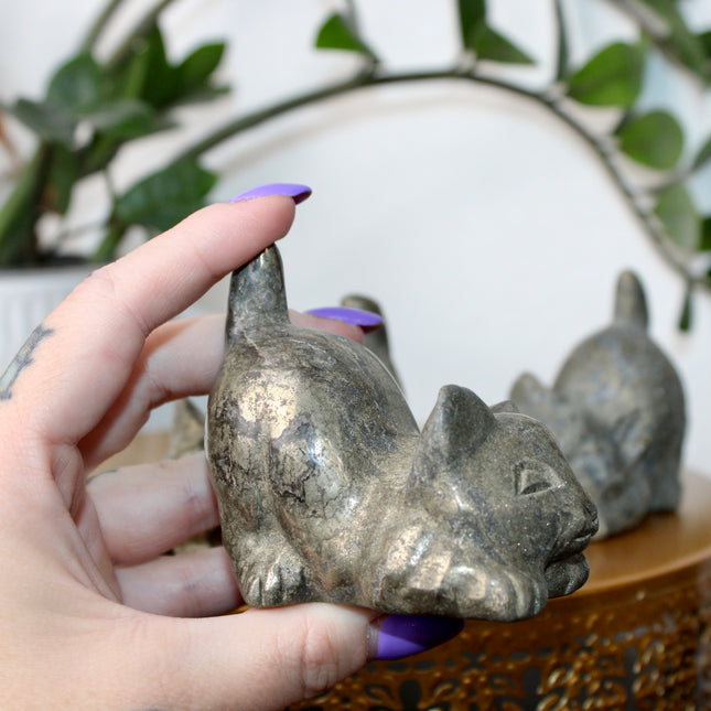 Hand holding a small stone Pyrite kitty cat carving another similar figurine in the background.