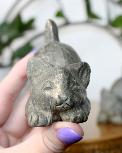 Hand holding a small stone Pyrite kitty cat carving another similar figurine in the background.