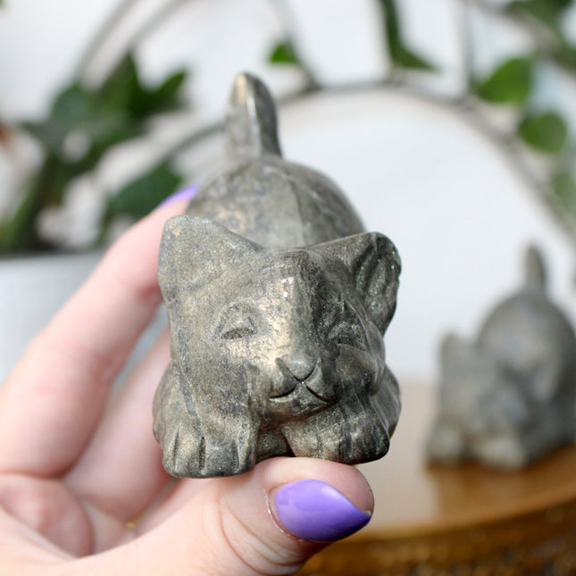 Hand holding a small stone Pyrite kitty cat carving another similar figurine in the background.
