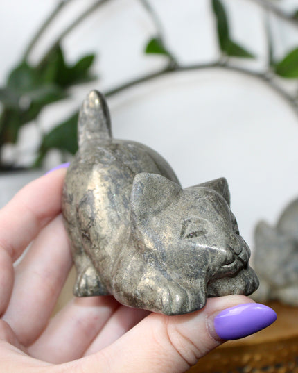 Hand holding a small stone Pyrite kitty cat carving another similar figurine in the background.
