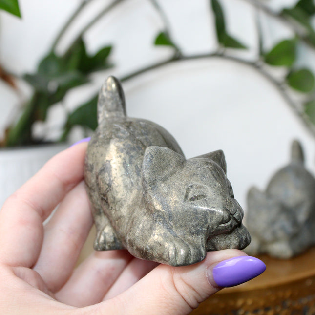 Hand holding a small stone Pyrite kitty cat carving another similar figurine in the background.