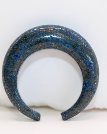 A striking Genuine Lapis Lazuli Stone Septum Gauge rests gently against a neutral-toned surface. The deep royal blue of the stone is rich and captivating, marbled with subtle veins of white calcite and scattered golden flecks of pyrite—classic hallmarks of authentic lapis. Expertly carved and polished into a smooth, curved shape for septum wear, the piece blends bold elegance with earthy mystique. Its surface gleams under soft lighting, showcasing the depth and natural beauty of the stone