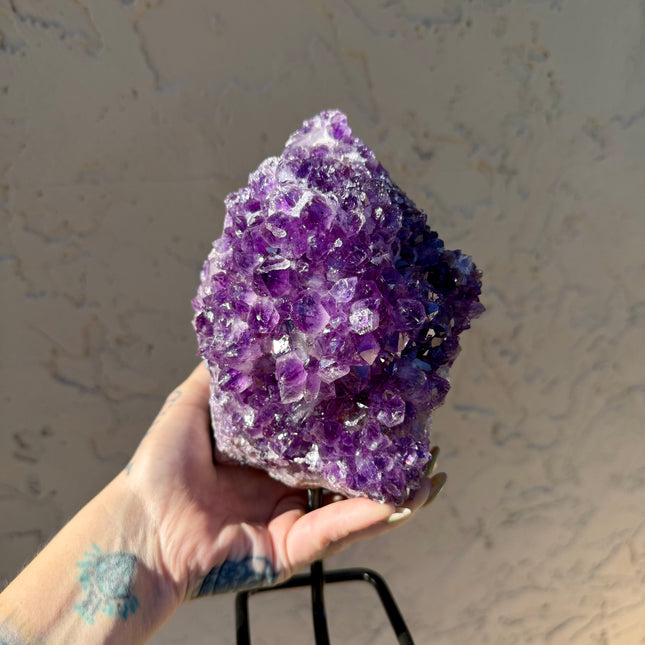 A dazzling Genuine Grade A Amethyst Druzy Geode rests on a natural surface, shimmering with tightly clustered crystal points in deep, vibrant purple. The interior of the geode sparkles with fine druzy crystals, catching the light like sugar-coated gemstones. This high-grade piece showcases rich violet hues, excellent clarity, and a dense crystal formation—hallmarks of premium quality amethyst. The outer shell maintains its raw, earthy texture, creating a striking contrast to the radiant interior. Perfect fo