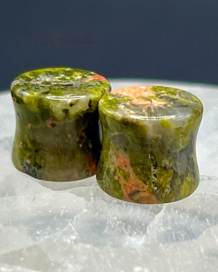 A grounded and vibrant pair of Unakite Double Flare Stone Plugs is displayed on a textured natural background. Each plug showcases the stone’s distinctive blend of mossy green epidote and soft, salmon-pink feldspar—creating a one-of-a-kind marbled pattern that feels both earthy and energizing. The smooth, hand-polished finish enhances the natural contrast in the stone’s texture and color. With a double flare shape for secure, comfortable wear, these plugs combine style with the natural, balancing energy Una