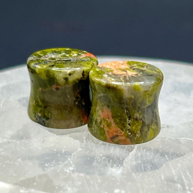 A grounded and vibrant pair of Unakite Double Flare Stone Plugs is displayed on a textured natural background. Each plug showcases the stone’s distinctive blend of mossy green epidote and soft, salmon-pink feldspar—creating a one-of-a-kind marbled pattern that feels both earthy and energizing. The smooth, hand-polished finish enhances the natural contrast in the stone’s texture and color. With a double flare shape for secure, comfortable wear, these plugs combine style with the natural, balancing energy Una