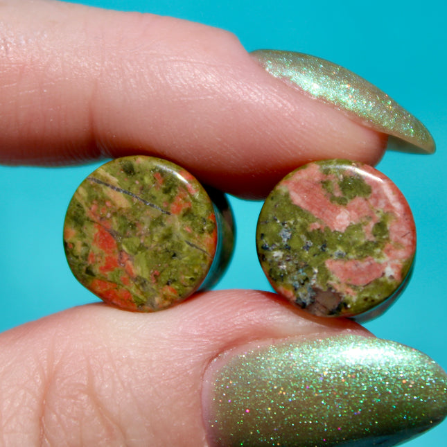 A grounded and vibrant pair of Unakite Double Flare Stone Plugs is displayed on a textured natural background. Each plug showcases the stone’s distinctive blend of mossy green epidote and soft, salmon-pink feldspar—creating a one-of-a-kind marbled pattern that feels both earthy and energizing. The smooth, hand-polished finish enhances the natural contrast in the stone’s texture and color. With a double flare shape for secure, comfortable wear, these plugs combine style with the natural, balancing energy Una