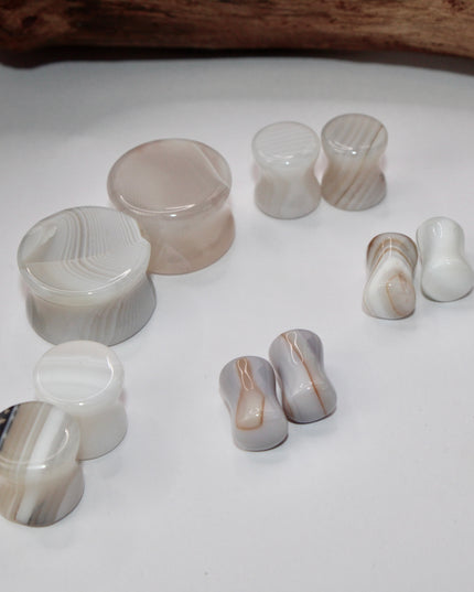 A serene pair of White Line Agate Plugs rests on a soft, neutral background, showcasing their natural, minimalist beauty. Each plug features delicate layers of white, cream, and translucent tones, with subtle linear banding unique to every piece. The polished finish enhances the stone’s soft sheen and calming aesthetic.