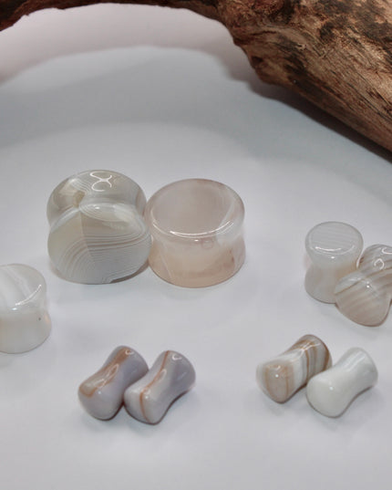 A serene pair of White Line Agate Plugs rests on a soft, neutral background, showcasing their natural, minimalist beauty. Each plug features delicate layers of white, cream, and translucent tones, with subtle linear banding unique to every piece. The polished finish enhances the stone’s soft sheen and calming aesthetic.