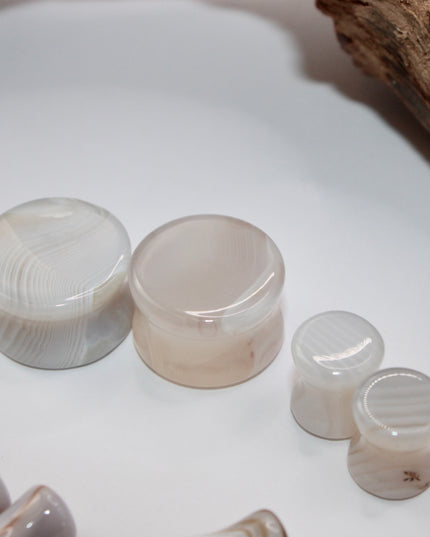 A serene pair of White Line Agate Plugs rests on a soft, neutral background, showcasing their natural, minimalist beauty. Each plug features delicate layers of white, cream, and translucent tones, with subtle linear banding unique to every piece. The polished finish enhances the stone’s soft sheen and calming aesthetic.