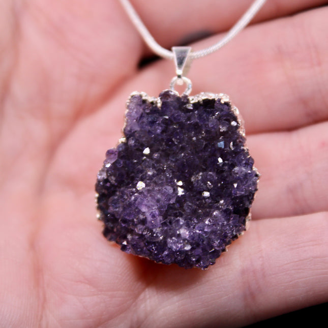 A beautiful Amethyst Pendant hangs gracefully from a sleek silver-color chain, combining natural elegance with everyday wearability. The pendant features a polished amethyst point or cabochon (style can vary), showcasing rich purple hues with subtle marbling or translucence that catches the light. Set in a simple metal cap or loop, the natural stone takes center stage. The silver-tone chain adds a modern, minimalist touch and pairs effortlessly with any outfit.