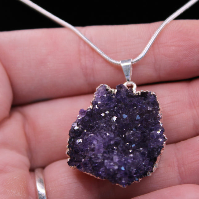 A beautiful Amethyst Pendant hangs gracefully from a sleek silver-color chain, combining natural elegance with everyday wearability. The pendant features a polished amethyst point or cabochon (style can vary), showcasing rich purple hues with subtle marbling or translucence that catches the light. Set in a simple metal cap or loop, the natural stone takes center stage. The silver-tone chain adds a modern, minimalist touch and pairs effortlessly with any outfit.