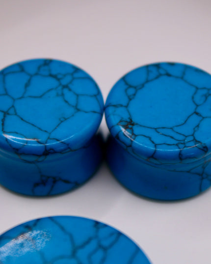 A vibrant pair of Blue Howlite Stone Plugs is shown resting on a soft beige surface. The plugs are round, polished to a high shine, and display a brilliant turquoise-blue color. Each plug is marked with distinctive dark gray to black veining in a web-like pattern—natural to howlite that has been dyed blue to resemble turquoise. The smooth, glossy finish enhances the contrast between the bright blue background and the delicate marbling. The lighting gently reflects off the curved surfaces, emphasizing their