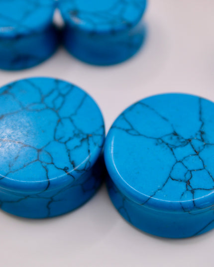 A vibrant pair of Blue Howlite Stone Plugs is shown resting on a soft beige surface. The plugs are round, polished to a high shine, and display a brilliant turquoise-blue color. Each plug is marked with distinctive dark gray to black veining in a web-like pattern—natural to howlite that has been dyed blue to resemble turquoise. The smooth, glossy finish enhances the contrast between the bright blue background and the delicate marbling. The lighting gently reflects off the curved surfaces, emphasizing their