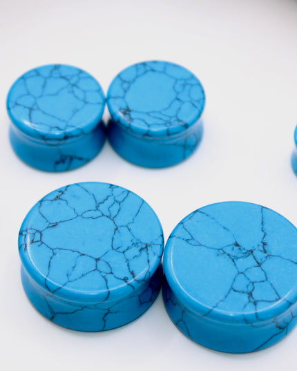 A vibrant pair of Blue Howlite Stone Plugs is shown resting on a soft beige surface. The plugs are round, polished to a high shine, and display a brilliant turquoise-blue color. Each plug is marked with distinctive dark gray to black veining in a web-like pattern—natural to howlite that has been dyed blue to resemble turquoise. The smooth, glossy finish enhances the contrast between the bright blue background and the delicate marbling. The lighting gently reflects off the curved surfaces, emphasizing their