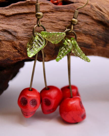 A close-up of a pair of striking Gothic earrings featuring glossy, deep-red cherry charms hanging from small silver skulls. The cherries appear juicy and vibrant, with a subtle dark shine, while the skulls add a bold, edgy contrast. The earrings are displayed against a black velvet background, enhancing their rich colors and alt-aesthetic vibe. Perfect for alternative, punk, or gothic fashion lovers who enjoy mixing sweetness with a hint of rebellion.