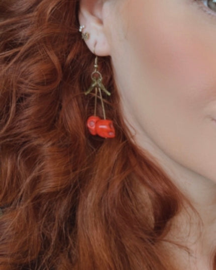 A close-up of a pair of striking Gothic earrings featuring glossy, deep-red cherry charms hanging from small silver skulls. The cherries appear juicy and vibrant, with a subtle dark shine, while the skulls add a bold, edgy contrast. The earrings are displayed against a black velvet background, enhancing their rich colors and alt-aesthetic vibe. Perfect for alternative, punk, or gothic fashion lovers who enjoy mixing sweetness with a hint of rebellion.