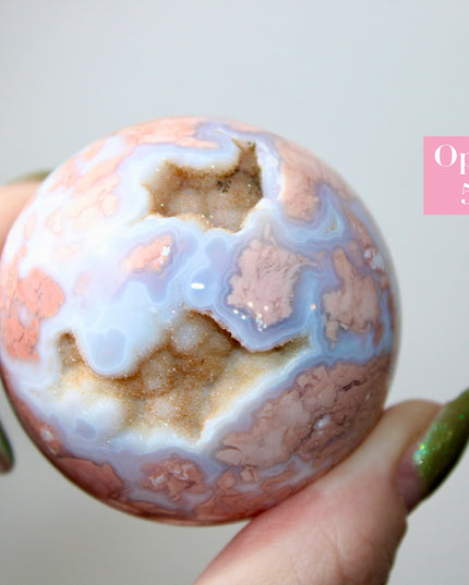 Elevate your space with the calming beauty and gentle energy of this hand-polished Flower Agate Sphere, a natural work of art that inspires peace, growth, and intention. Featuring dreamy shades of soft pink, creamy white, and warm peach, each sphere showcases unique floral-like inclusions that resemble delicate blooms suspended within the stone—making every piece truly one of a kind.