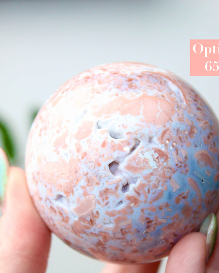Elevate your space with the calming beauty and gentle energy of this hand-polished Flower Agate Sphere, a natural work of art that inspires peace, growth, and intention. Featuring dreamy shades of soft pink, creamy white, and warm peach, each sphere showcases unique floral-like inclusions that resemble delicate blooms suspended within the stone—making every piece truly one of a kind.