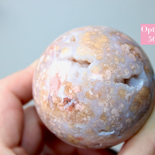 Elevate your space with the calming beauty and gentle energy of this hand-polished Flower Agate Sphere, a natural work of art that inspires peace, growth, and intention. Featuring dreamy shades of soft pink, creamy white, and warm peach, each sphere showcases unique floral-like inclusions that resemble delicate blooms suspended within the stone—making every piece truly one of a kind.