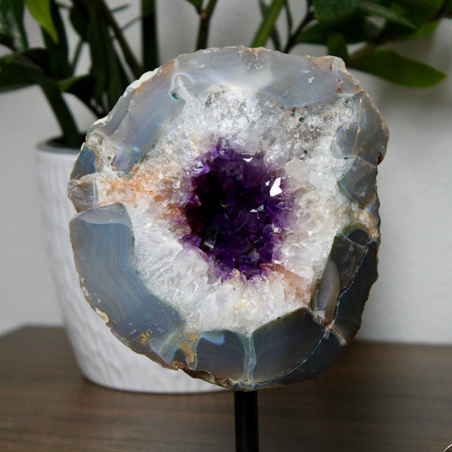 An impressive Large Geode stands tall on its included display stand, revealing a breathtaking Grade A Dark Purple Amethyst Druzy Center. The interior is densely lined with sparkling amethyst crystals, rich in deep violet tones and glowing with natural brilliance. The druzy surface catches the light with every angle, creating a glittering effect that highlights the exceptional clarity and saturation of the crystals. The raw outer shell offers a rugged, earthy contrast to the radiant core, emphasizing the geo