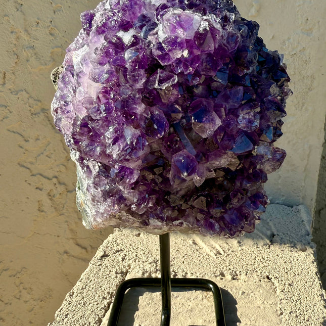 A dazzling Genuine Grade A Amethyst Druzy Geode rests on a natural surface, shimmering with tightly clustered crystal points in deep, vibrant purple. The interior of the geode sparkles with fine druzy crystals, catching the light like sugar-coated gemstones. This high-grade piece showcases rich violet hues, excellent clarity, and a dense crystal formation—hallmarks of premium quality amethyst. The outer shell maintains its raw, earthy texture, creating a striking contrast to the radiant interior. Perfect fo