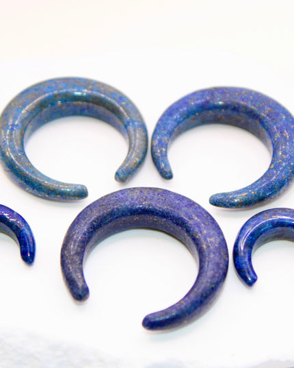 A striking Genuine Lapis Lazuli Stone Septum Gauge rests gently against a neutral-toned surface. The deep royal blue of the stone is rich and captivating, marbled with subtle veins of white calcite and scattered golden flecks of pyrite—classic hallmarks of authentic lapis. Expertly carved and polished into a smooth, curved shape for septum wear, the piece blends bold elegance with earthy mystique. Its surface gleams under soft lighting, showcasing the depth and natural beauty of the stone