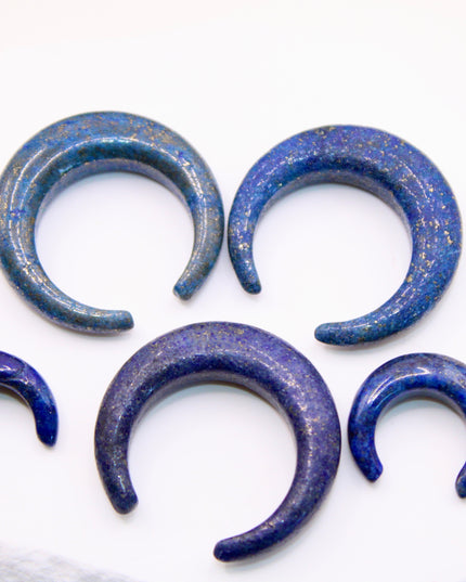 A striking Genuine Lapis Lazuli Stone Septum Gauge rests gently against a neutral-toned surface. The deep royal blue of the stone is rich and captivating, marbled with subtle veins of white calcite and scattered golden flecks of pyrite—classic hallmarks of authentic lapis. Expertly carved and polished into a smooth, curved shape for septum wear, the piece blends bold elegance with earthy mystique. Its surface gleams under soft lighting, showcasing the depth and natural beauty of the stone