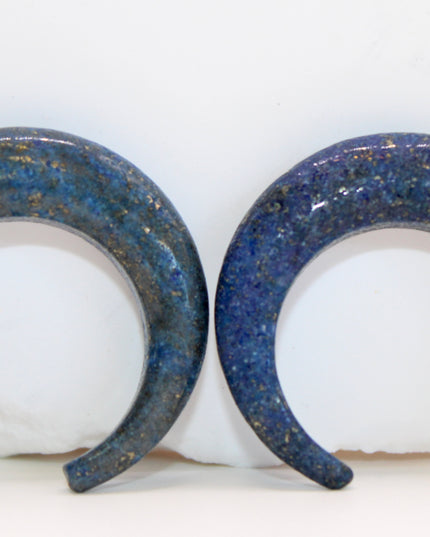 A striking Genuine Lapis Lazuli Stone Septum Gauge rests gently against a neutral-toned surface. The deep royal blue of the stone is rich and captivating, marbled with subtle veins of white calcite and scattered golden flecks of pyrite—classic hallmarks of authentic lapis. Expertly carved and polished into a smooth, curved shape for septum wear, the piece blends bold elegance with earthy mystique. Its surface gleams under soft lighting, showcasing the depth and natural beauty of the stone