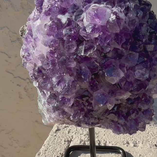 A dazzling Genuine Grade A Amethyst Druzy Geode rests on a natural surface, shimmering with tightly clustered crystal points in deep, vibrant purple. The interior of the geode sparkles with fine druzy crystals, catching the light like sugar-coated gemstones. This high-grade piece showcases rich violet hues, excellent clarity, and a dense crystal formation—hallmarks of premium quality amethyst. The outer shell maintains its raw, earthy texture, creating a striking contrast to the radiant interior. Perfect fo