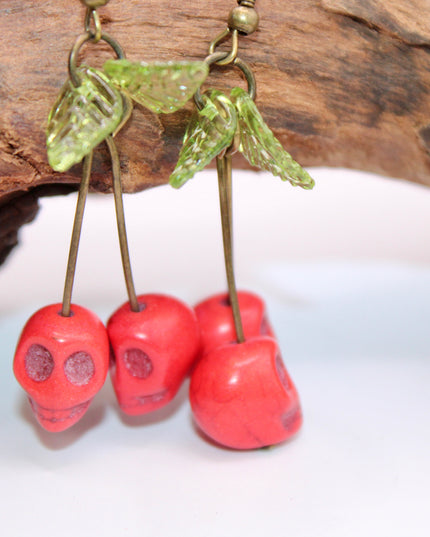 A close-up of a pair of striking Gothic earrings featuring glossy, deep-red cherry charms hanging from small silver skulls. The cherries appear juicy and vibrant, with a subtle dark shine, while the skulls add a bold, edgy contrast. The earrings are displayed against a black velvet background, enhancing their rich colors and alt-aesthetic vibe. Perfect for alternative, punk, or gothic fashion lovers who enjoy mixing sweetness with a hint of rebellion.