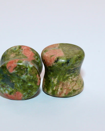 A grounded and vibrant pair of Unakite Double Flare Stone Plugs is displayed on a textured natural background. Each plug showcases the stone’s distinctive blend of mossy green epidote and soft, salmon-pink feldspar—creating a one-of-a-kind marbled pattern that feels both earthy and energizing. The smooth, hand-polished finish enhances the natural contrast in the stone’s texture and color. With a double flare shape for secure, comfortable wear, these plugs combine style with the natural, balancing energy