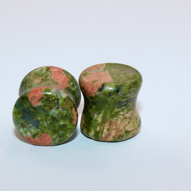 A grounded and vibrant pair of Unakite Double Flare Stone Plugs is displayed on a textured natural background. Each plug showcases the stone’s distinctive blend of mossy green epidote and soft, salmon-pink feldspar—creating a one-of-a-kind marbled pattern that feels both earthy and energizing. The smooth, hand-polished finish enhances the natural contrast in the stone’s texture and color. With a double flare shape for secure, comfortable wear, these plugs combine style with the natural, balancing energy