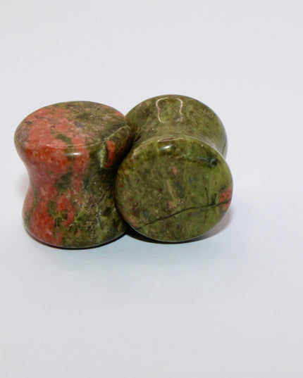 A grounded and vibrant pair of Unakite Double Flare Stone Plugs is displayed on a textured natural background. Each plug showcases the stone’s distinctive blend of mossy green epidote and soft, salmon-pink feldspar—creating a one-of-a-kind marbled pattern that feels both earthy and energizing. The smooth, hand-polished finish enhances the natural contrast in the stone’s texture and color. With a double flare shape for secure, comfortable wear, these plugs combine style with the natural, balancing energy Una