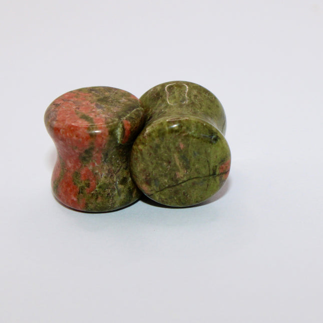 A grounded and vibrant pair of Unakite Double Flare Stone Plugs is displayed on a textured natural background. Each plug showcases the stone’s distinctive blend of mossy green epidote and soft, salmon-pink feldspar—creating a one-of-a-kind marbled pattern that feels both earthy and energizing. The smooth, hand-polished finish enhances the natural contrast in the stone’s texture and color. With a double flare shape for secure, comfortable wear, these plugs combine style with the natural, balancing energy Una
