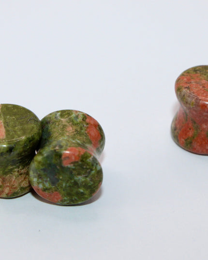 A grounded and vibrant pair of Unakite Double Flare Stone Plugs is displayed on a textured natural background. Each plug showcases the stone’s distinctive blend of mossy green epidote and soft, salmon-pink feldspar—creating a one-of-a-kind marbled pattern that feels both earthy and energizing. The smooth, hand-polished finish enhances the natural contrast in the stone’s texture and color. With a double flare shape for secure, comfortable wear, these plugs combine style with the natural, balancing energy Una