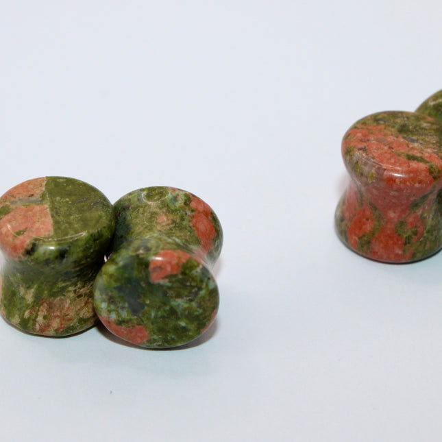 A grounded and vibrant pair of Unakite Double Flare Stone Plugs is displayed on a textured natural background. Each plug showcases the stone’s distinctive blend of mossy green epidote and soft, salmon-pink feldspar—creating a one-of-a-kind marbled pattern that feels both earthy and energizing. The smooth, hand-polished finish enhances the natural contrast in the stone’s texture and color. With a double flare shape for secure, comfortable wear, these plugs combine style with the natural, balancing energy Una