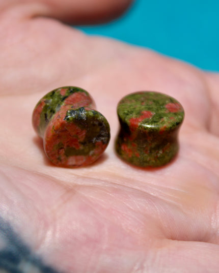 A grounded and vibrant pair of Unakite Double Flare Stone Plugs is displayed on a textured natural background. Each plug showcases the stone’s distinctive blend of mossy green epidote and soft, salmon-pink feldspar—creating a one-of-a-kind marbled pattern that feels both earthy and energizing. The smooth, hand-polished finish enhances the natural contrast in the stone’s texture and color. With a double flare shape for secure, comfortable wear, these plugs combine style with the natural, balancing energy Una