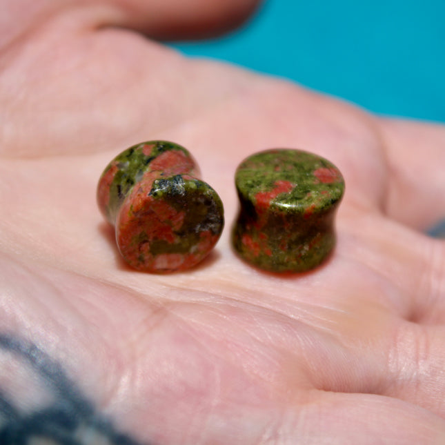 A grounded and vibrant pair of Unakite Double Flare Stone Plugs is displayed on a textured natural background. Each plug showcases the stone’s distinctive blend of mossy green epidote and soft, salmon-pink feldspar—creating a one-of-a-kind marbled pattern that feels both earthy and energizing. The smooth, hand-polished finish enhances the natural contrast in the stone’s texture and color. With a double flare shape for secure, comfortable wear, these plugs combine style with the natural, balancing energy Una